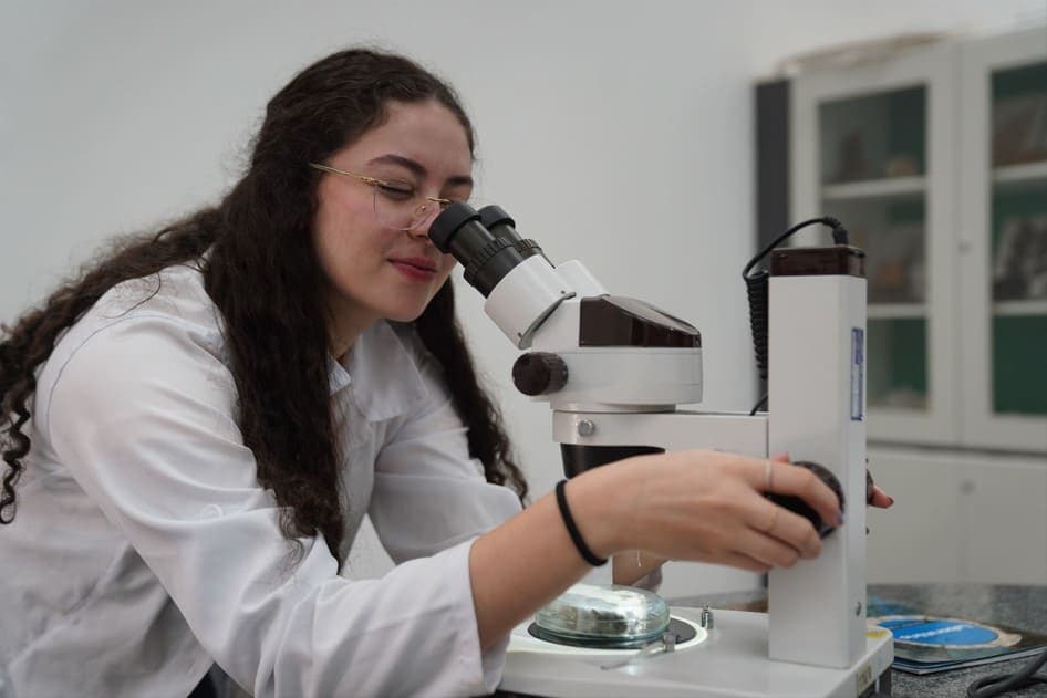 Uma estudante usando um microscópio em um laboratório, ajustando o equipamento com as mãos.