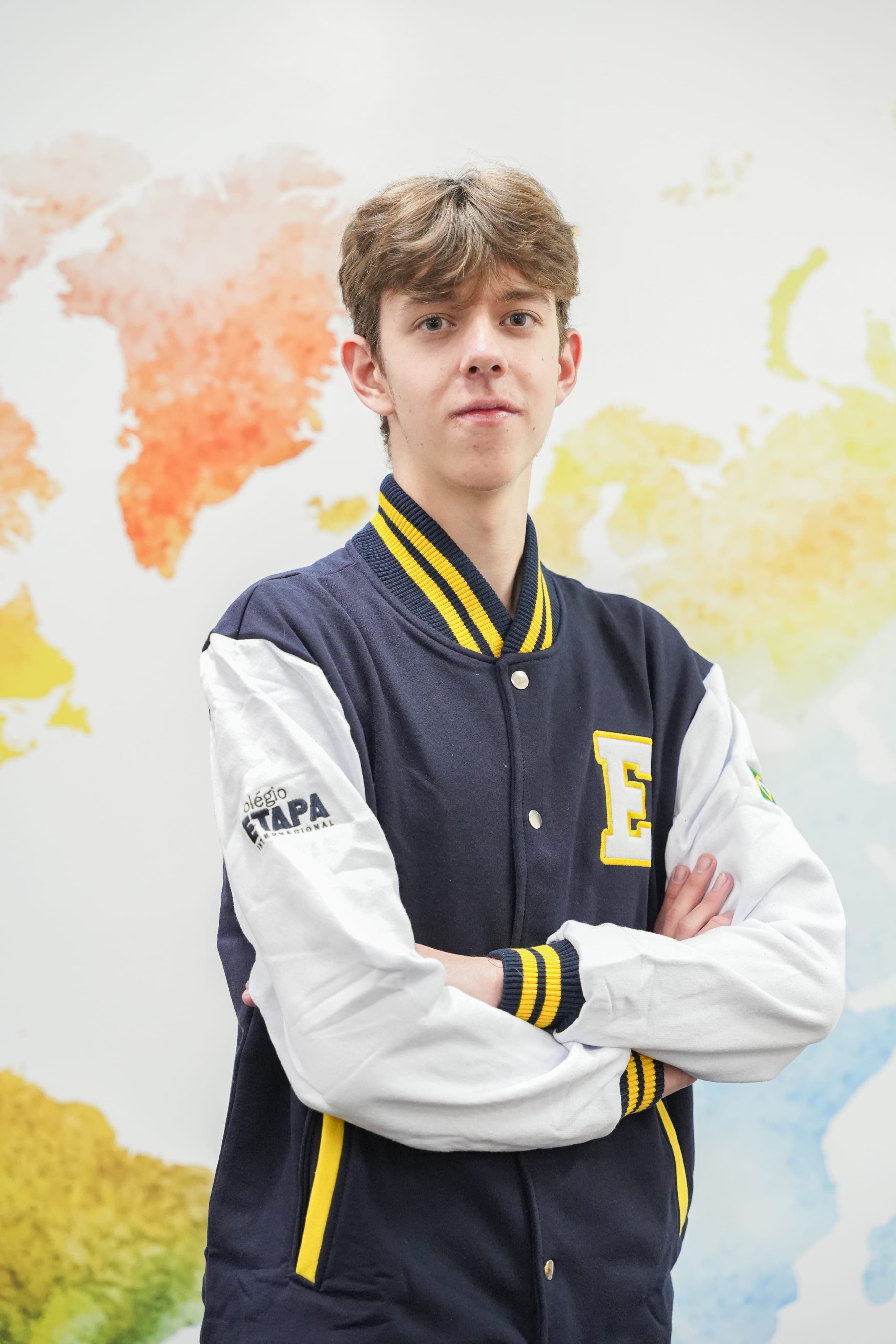 Um adolescente com os braços cruzados usando jaqueta escolar, em frente a um mapa-múndi colorido na parede.