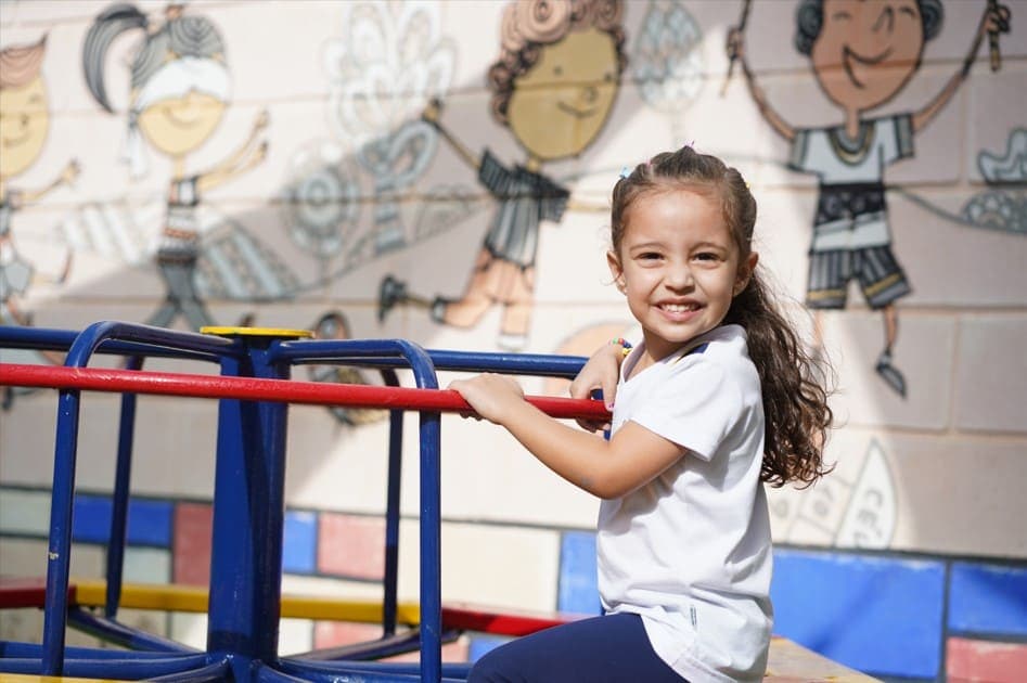 Uma menina sorrindo sentada em um brinquedo de playground, com mural de crianças desenhadas ao fundo.